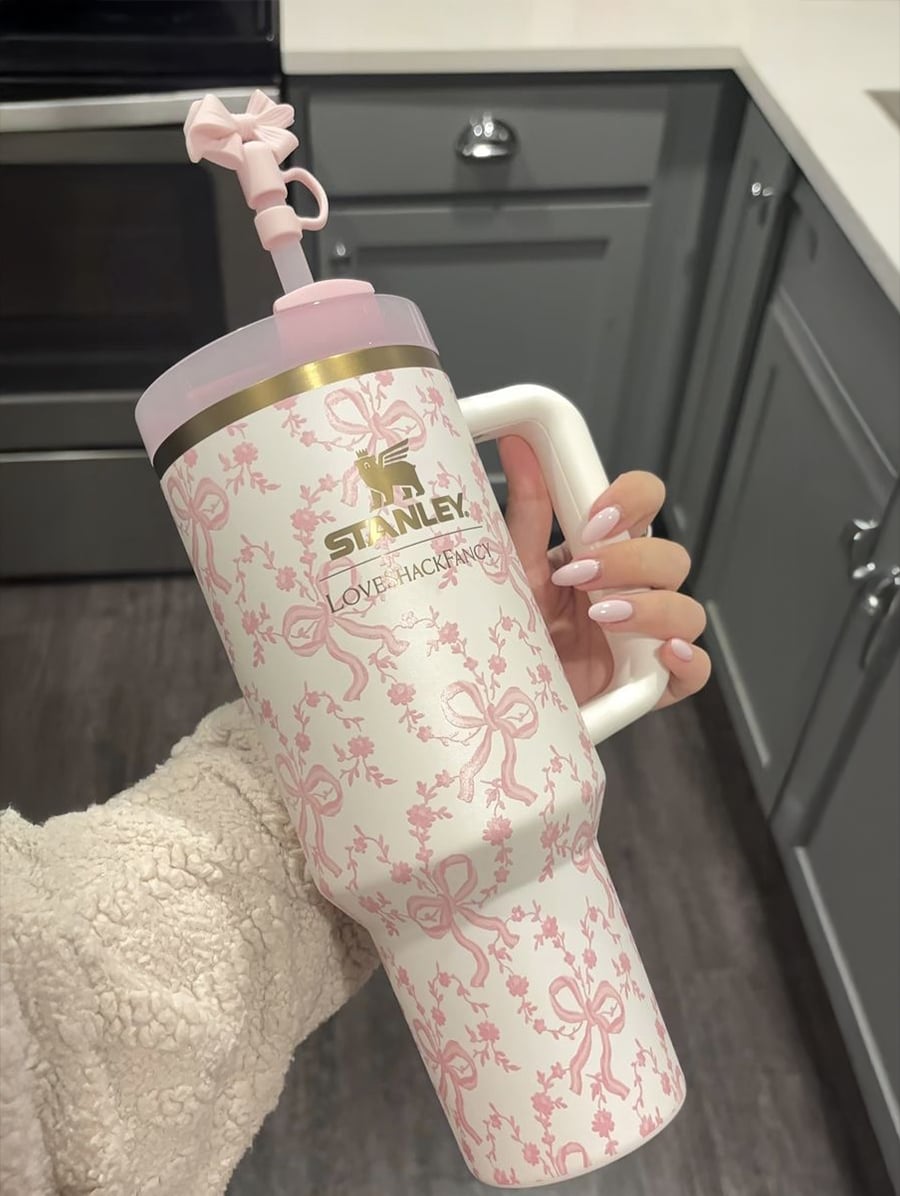 A hand holds a large Stanley tumbler with a pink bow pattern, matching straw topper, and white handle in a kitchen with gray cabinets.