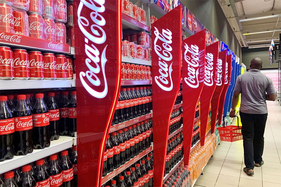 A man with a red basket walks past shelves stocked with various Coca-Cola bottles and cans in a brightly lit grocery store aisle.