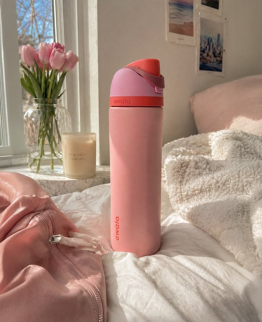 A pink Owala water bottle sits on a bed with cozy blankets near a window, with pink tulips, a candle, and wall photos in the background. Sunlight streams in, creating a warm, inviting atmosphere.