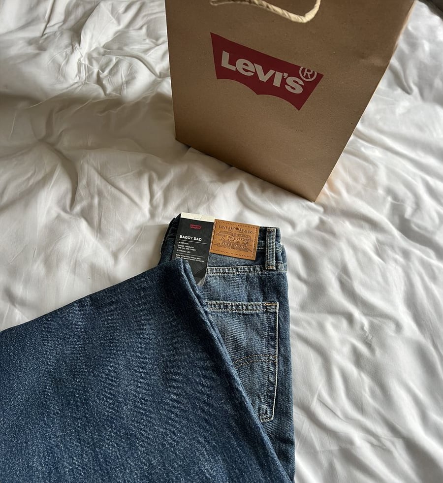A pair of folded Levi's blue jeans lies on a white bed next to a brown Levi's shopping bag.