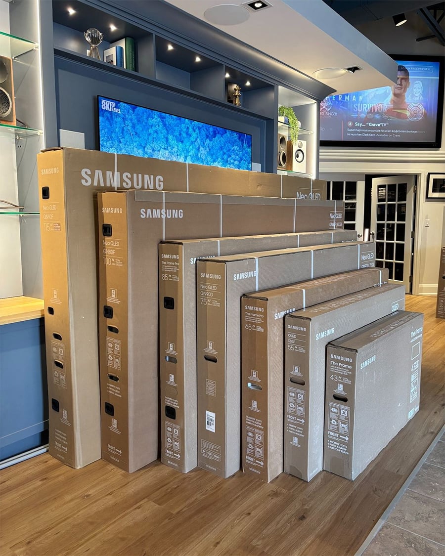 Several Samsung TV boxes of various sizes are lined up vertically inside a modern electronics store with wooden floors and display shelves.