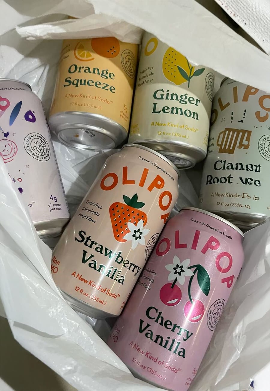 Six cans of OLIPOP soda in a white bag, featuring flavors Orange Squeeze, Ginger Lemon, Classic Root Beer, Strawberry Vanilla, and Cherry Vanilla. The cans are colorful and arranged closely together.
