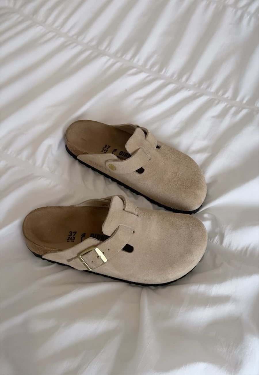 A pair of beige suede slip-on clogs with buckles rests on a white, wrinkled bedspread.