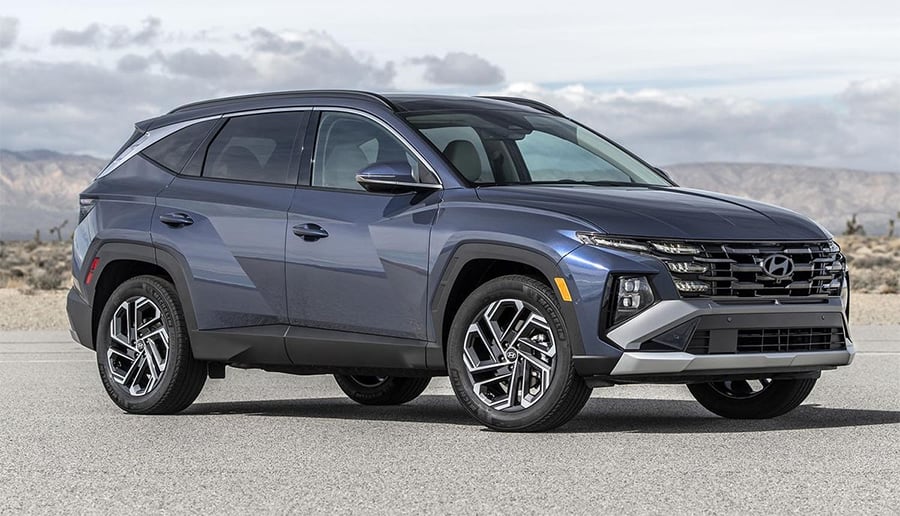 A blue Hyundai SUV is parked on a paved surface with mountains and a cloudy sky in the background.