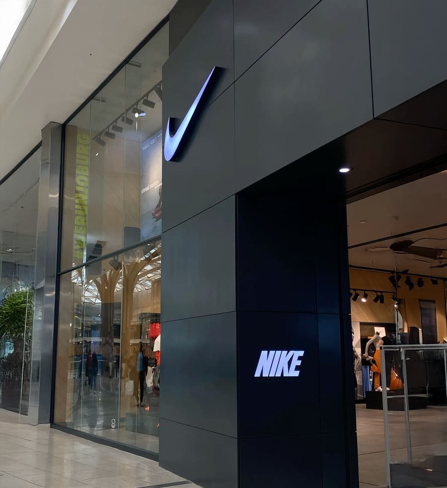 The entrance of a Nike store in a mall, featuring the Nike logo and brand name on a sleek black exterior panel near large glass windows.