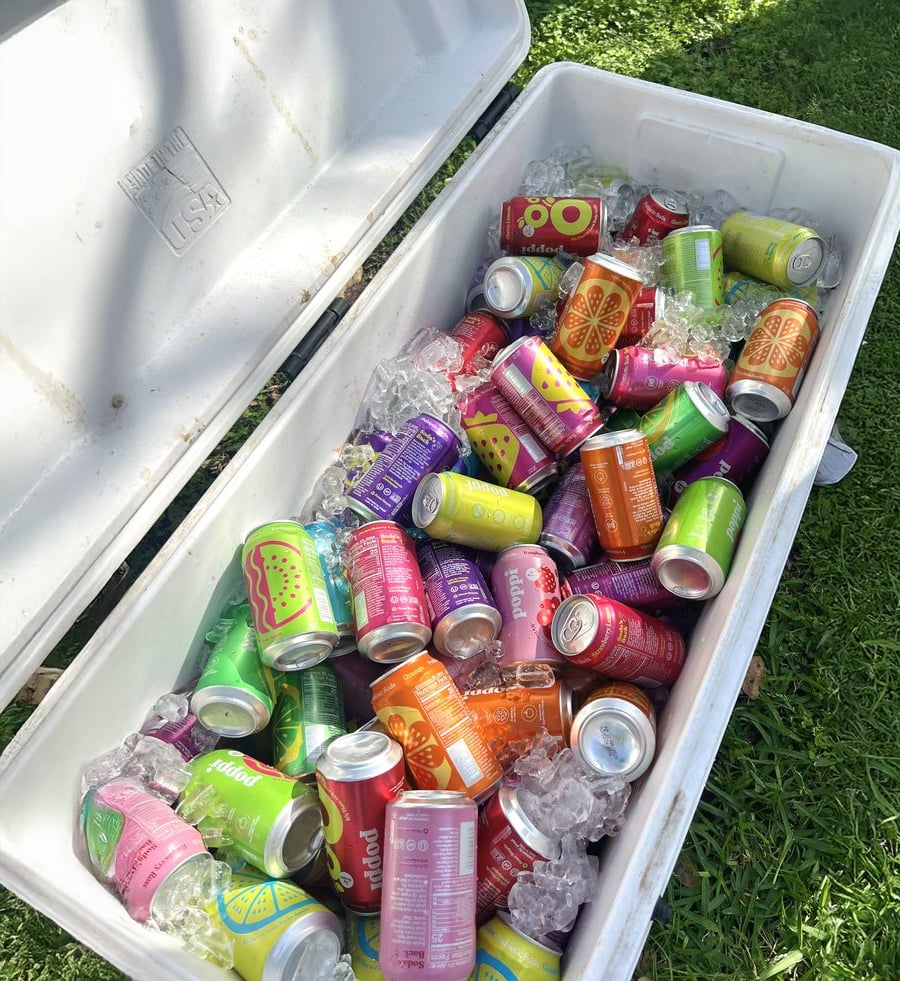 An open white cooler filled with colorful assorted soda cans and ice sits on green grass under sunlight.