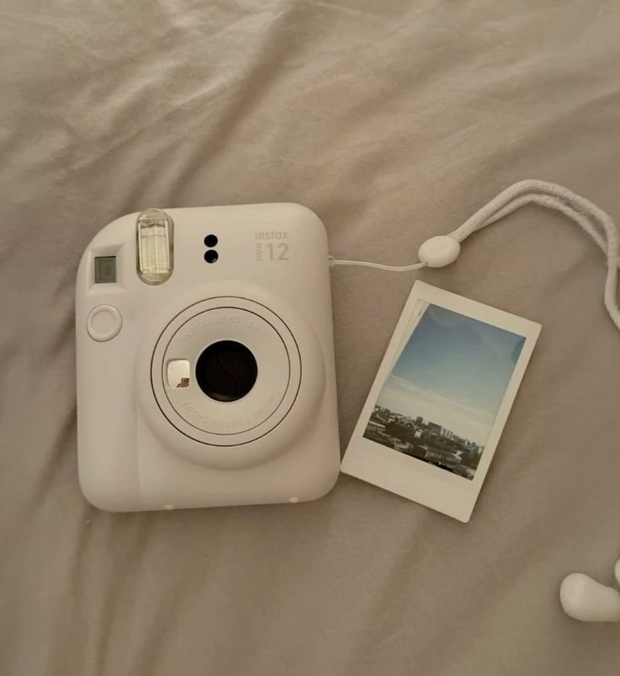 A white instant camera lies on a light bedspread beside a printed photo showing a cityscape. A strap is attached to the camera, and part of a white earbud is visible in the lower corner.