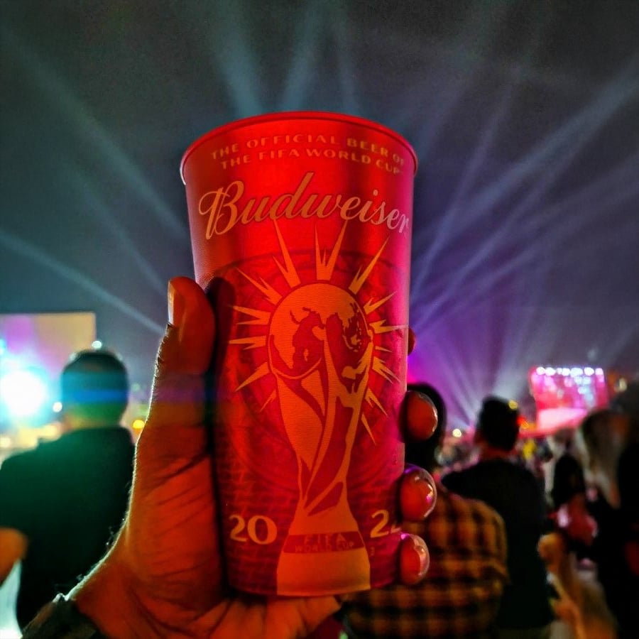 A hand holds a red Budweiser cup with a FIFA World Cup design at a night event, with colorful stage lights and a crowd in the background.