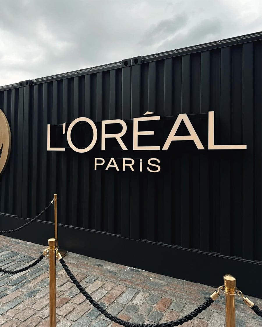 A large L'Oréal Paris sign is displayed on a black wall with gold lettering. A rope barrier is set up in front on a cobblestone pavement under a cloudy sky.