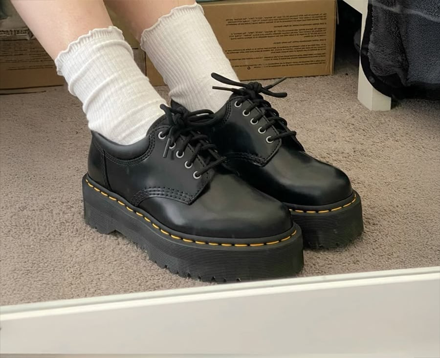 A person wearing white socks and black platform lace-up shoes stands on a beige carpet, with cardboard boxes and a mirror visible in the background.