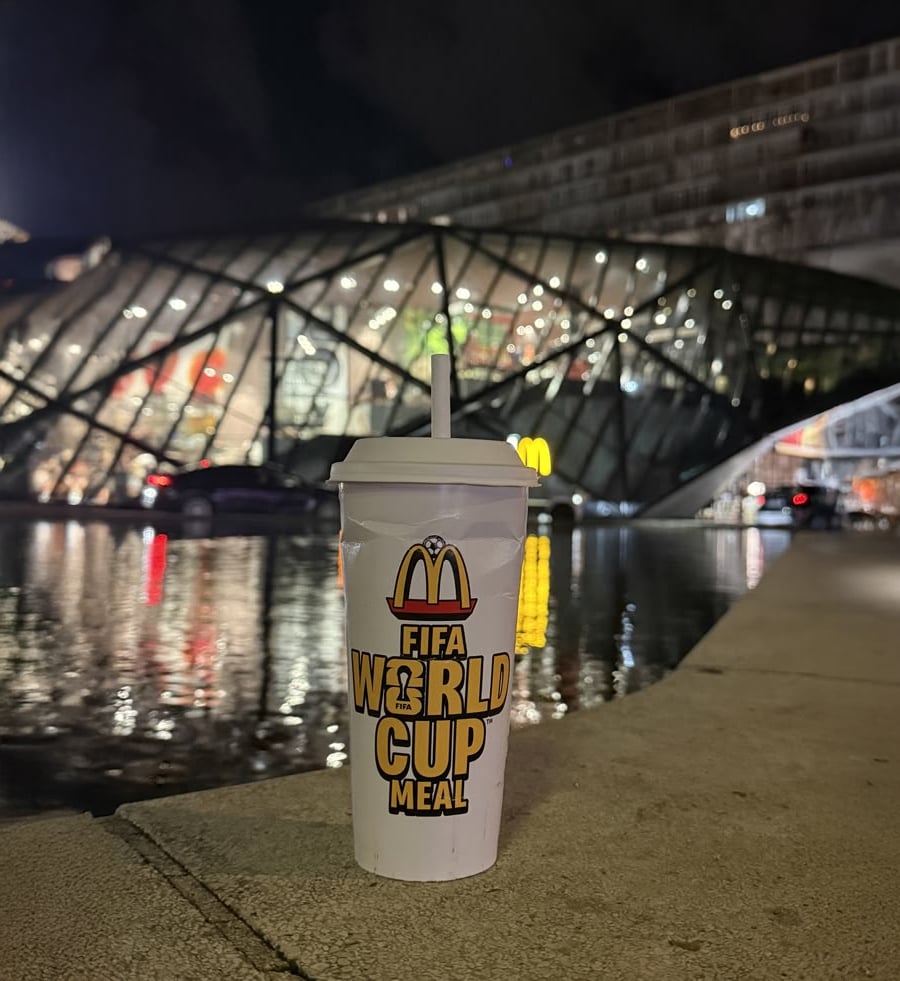 A McDonald’s FIFA World Cup Meal cup stands in front of a modern glass building at night, with reflections visible in nearby water and blurred lights in the background.