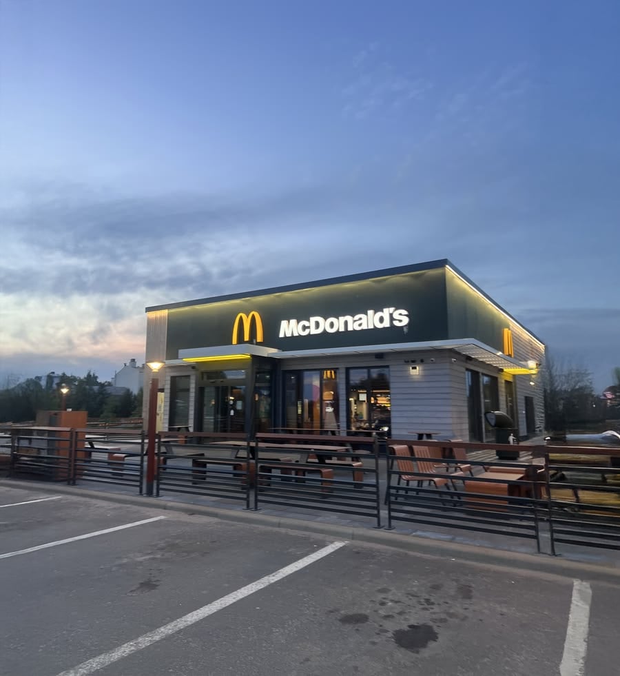 A McDonald's restaurant at dusk with illuminated signage, outdoor seating, and an empty parking lot in the foreground.