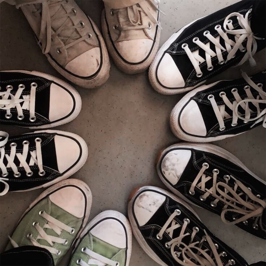 Eight pairs of sneakers, mostly black and white with one green pair, are arranged in a circle with toes pointing inward on a light-colored floor.