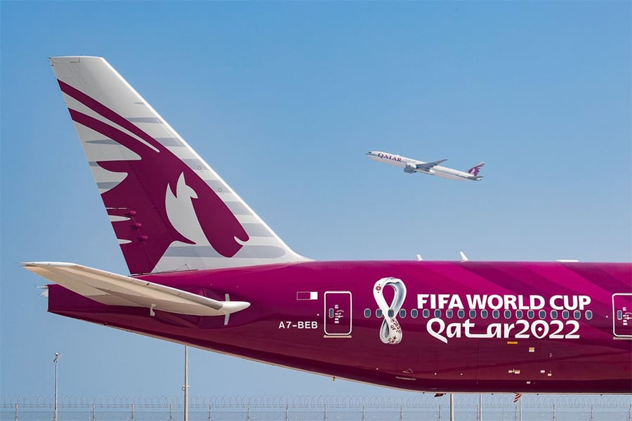 A Qatar Airways plane with FIFA World Cup Qatar 2022 branding is parked, while another plane takes off in the clear blue sky in the background.