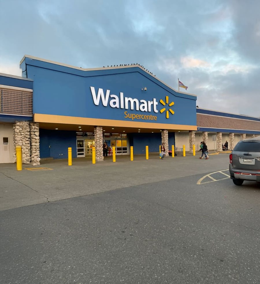 Walmart Supercentre exterior with blue and yellow signage, people entering and exiting, cars parked nearby, and a partly cloudy sky overhead.