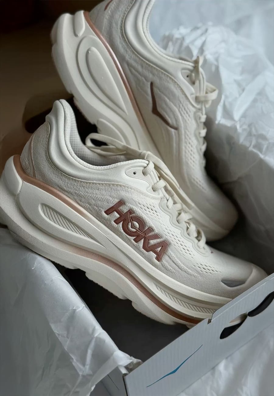 A pair of beige HOKA running shoes with thick soles is displayed in an open shoebox lined with white tissue paper.