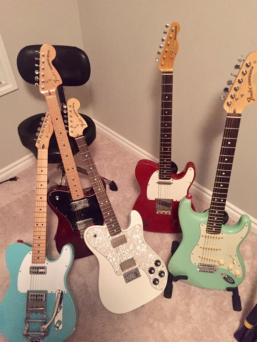 Four electric guitars in different colors (blue, white, red, and green) are positioned around a black chair on a carpeted floor in a corner of a room with beige walls.