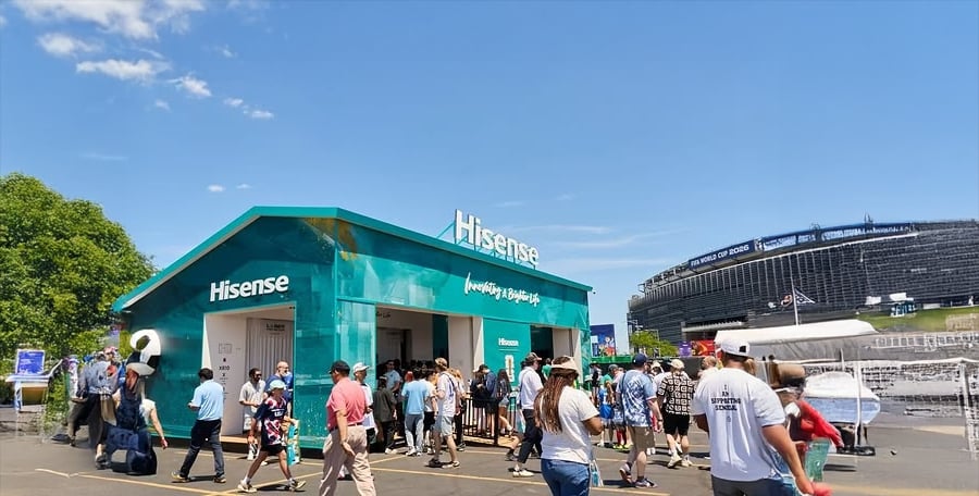 A crowd gathers outside a turquoise Hisense booth on a sunny day, with a stadium visible in the background and people walking around the event area.