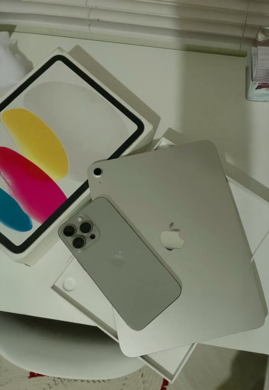 A silver iPhone and iPad rest on a table next to an open box with colorful oval inserts in yellow, pink, and blue. Blinds and part of a tissue box are visible in the background.