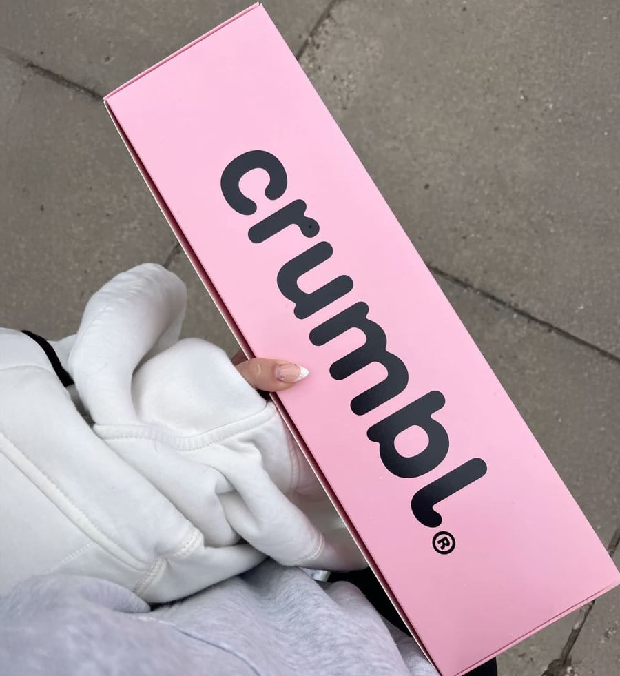 A person wearing a white hoodie holds a long pink Crumbl Cookies box against a concrete sidewalk.