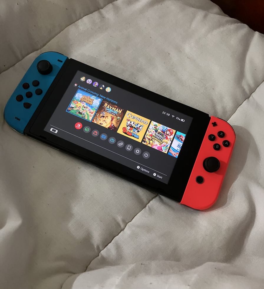 A Nintendo Switch with blue and red Joy-Con controllers rests on a white blanket, displaying the home screen with various game icons visible.