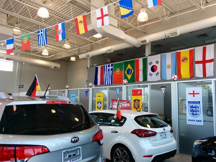 Cars inside a dealership decorated with international flags, including England, Brazil, Argentina, Germany, and more, likely celebrating a global sports event. The cars are silver and white, and soccer balls hang from the ceiling.