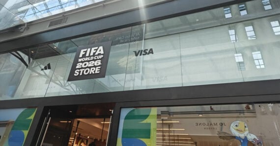 Glass storefront with "FIFA World Cup 2026 Store" sign, logos for adidas and VISA, and part of a colorful mascot mural visible on the lower windows. Reflections show ceiling beams and building structure above.