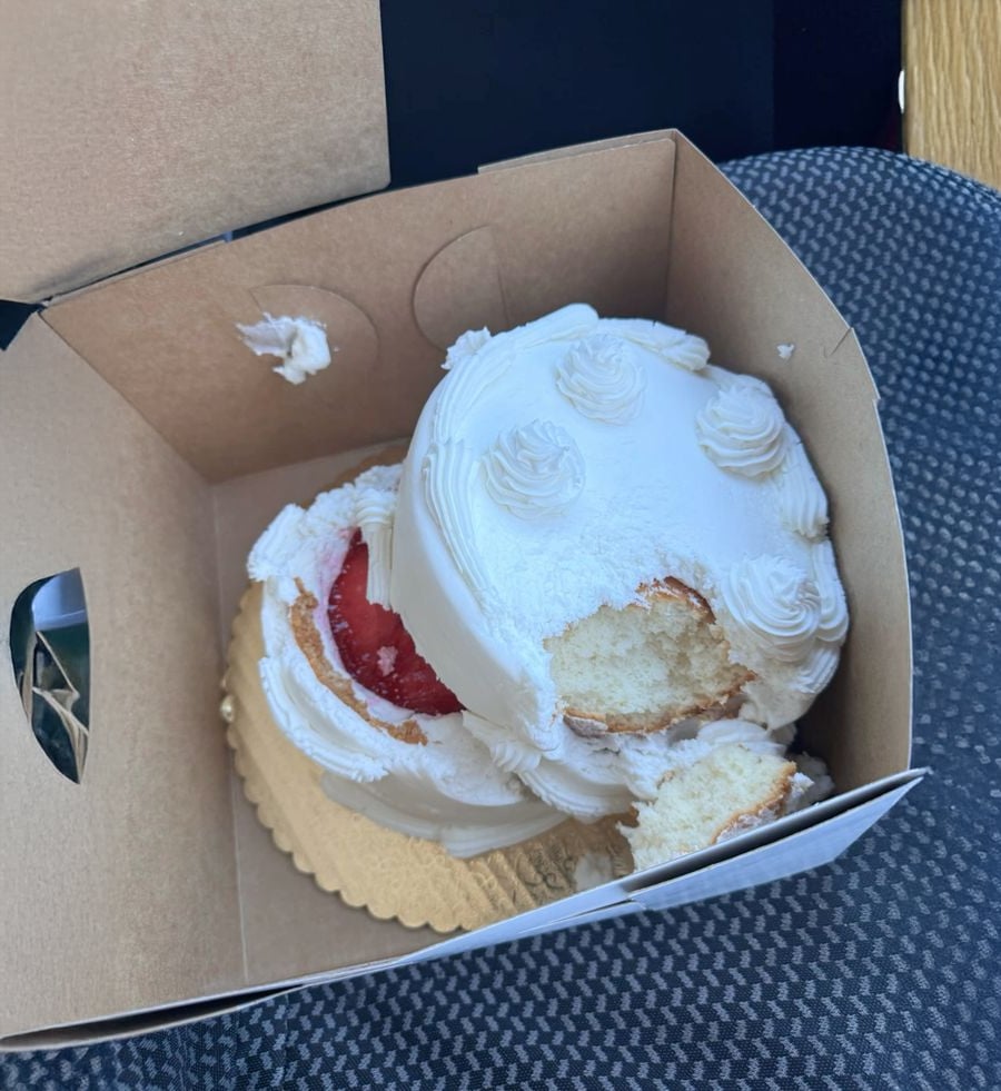A small, white-frosted cake with a strawberry center in a brown bakery box, missing a chunk as if bitten into, sits on a car seat.