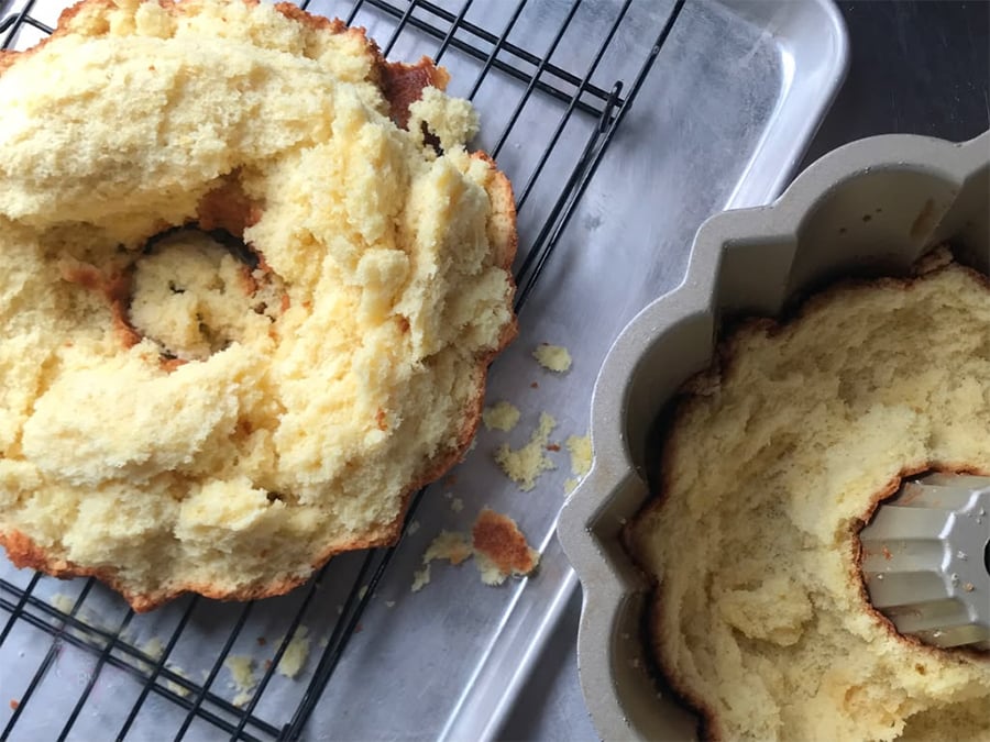 Broken bundt cake on a cooling rack with crumbs scattered nearby, and the remaining half still in the bundt pan, all placed on a metal baking tray.