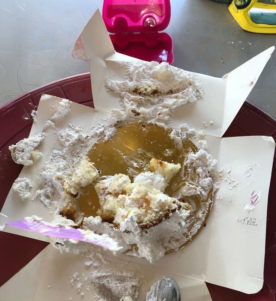 A partially eaten cake sits on a torn-open cake box, with crumbs and frosting scattered around. A spoon and a pink plastic container are nearby on a round table.