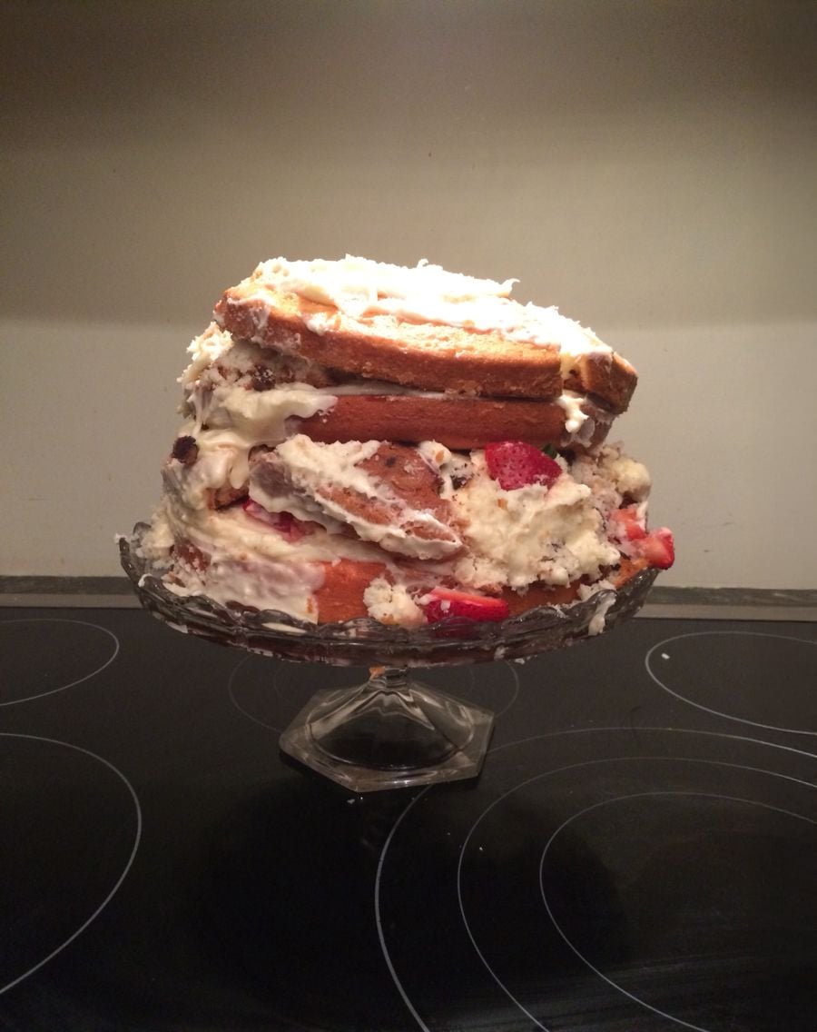 A messy, unevenly stacked layer cake with white frosting and pieces of strawberries, sitting on a glass cake stand on a stovetop.