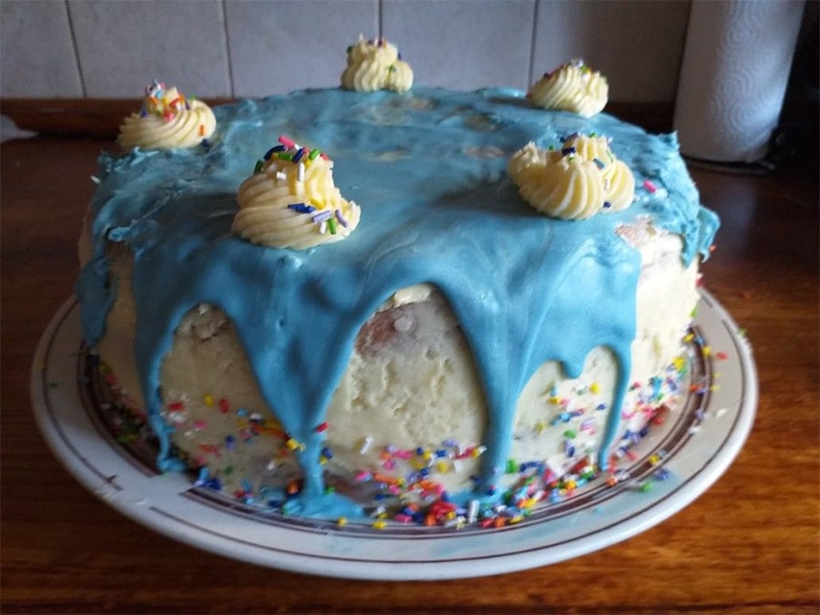 A round white cake with blue icing dripping over the edges, topped with five swirls of yellow frosting and colorful sprinkles on the plate and cake.