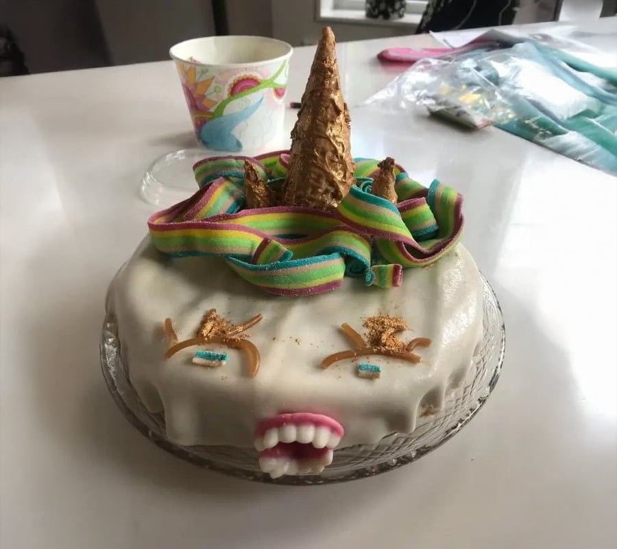 A homemade unicorn cake with a golden horn, rainbow candy mane, white icing, and funny fondant teeth, sitting on a glass plate on a white table.