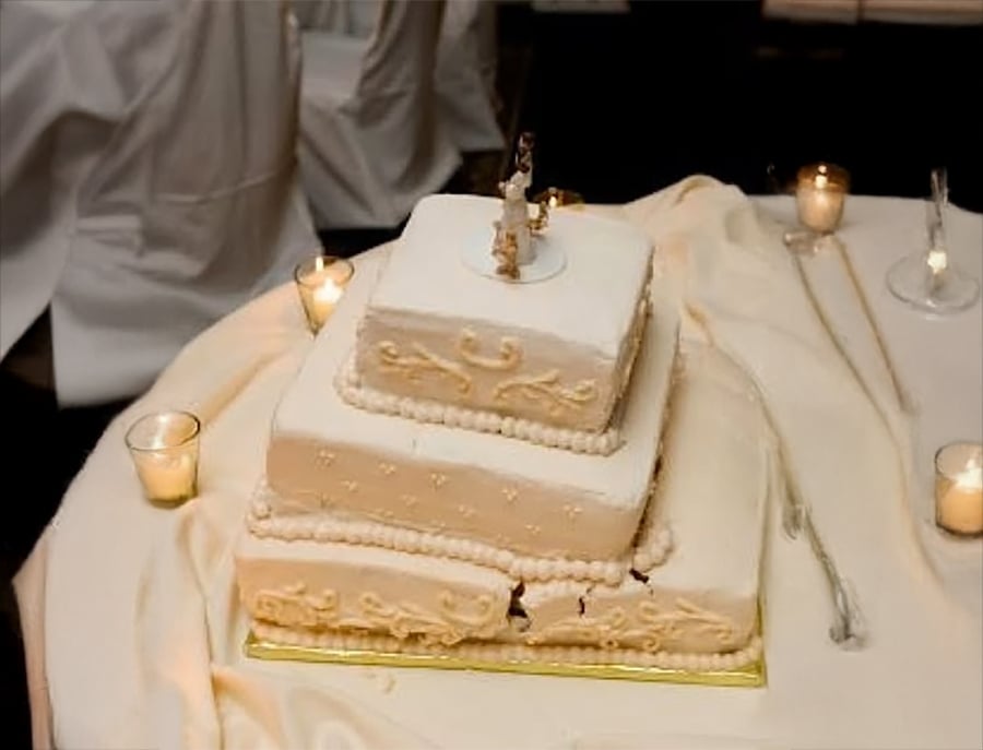 Three-tiered white wedding cake with decorative icing, some slices missing from the bottom tier, surrounded by lit candles on a cream-colored tablecloth.