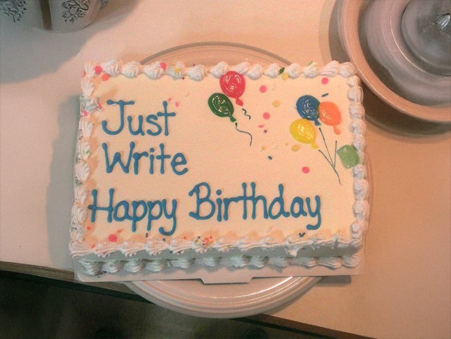 A rectangular birthday cake with white icing, balloon decorations, and the words "Just Write Happy Birthday" piped in blue frosting.