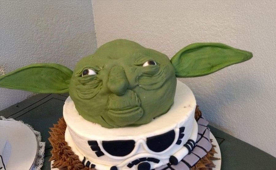 A two-tier cake features a green Yoda head with large ears on top, and Star Wars-themed decorations below, including a white band with black patterns resembling Chewbacca’s sash.