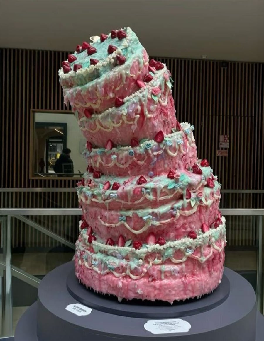 A large, colorful, leaning cake with pink, blue, and white icing, decorated with strawberries, sits on display indoors against a background of striped wooden wall panels.