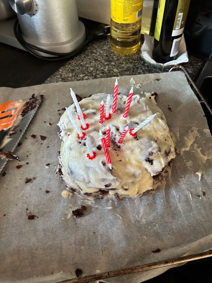 A homemade, square chocolate cake with uneven white icing, red and white candles, on parchment paper. A knife and spatula are beside the cake, with cooking oil bottles visible in the background.