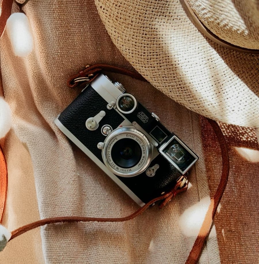 A vintage film camera rests on a beige fabric surface next to a woven straw hat with a brown leather strap. Sunlight creates soft, dappled patterns across the scene.