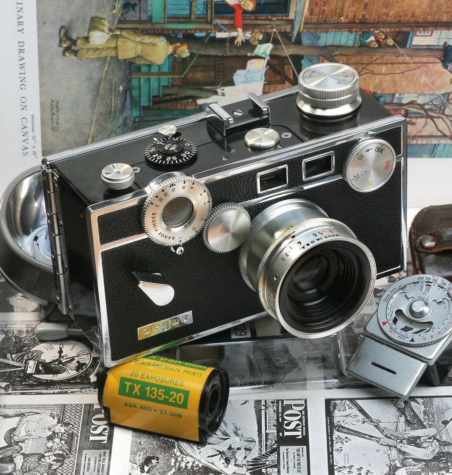 A vintage film camera with a light meter, film canister, and old photographs. The camera has several dials and a large lens, and the yellow film roll is labeled “TX 135-20.”