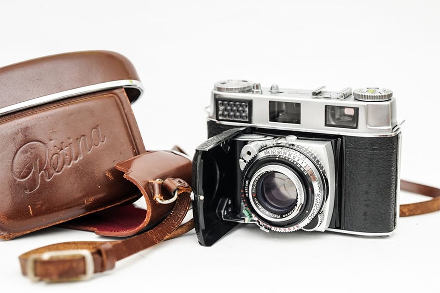 A vintage Kodak Retina camera with a leather case is placed on a white surface. The camera lens is open, and the brown case, embossed with "Retina," lies beside it with its strap visible.
