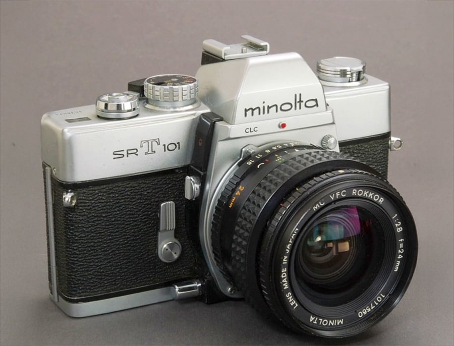 A vintage Minolta SR-T 101 film camera with a silver and black body, featuring a large lens and various dials, is placed on a plain gray surface.