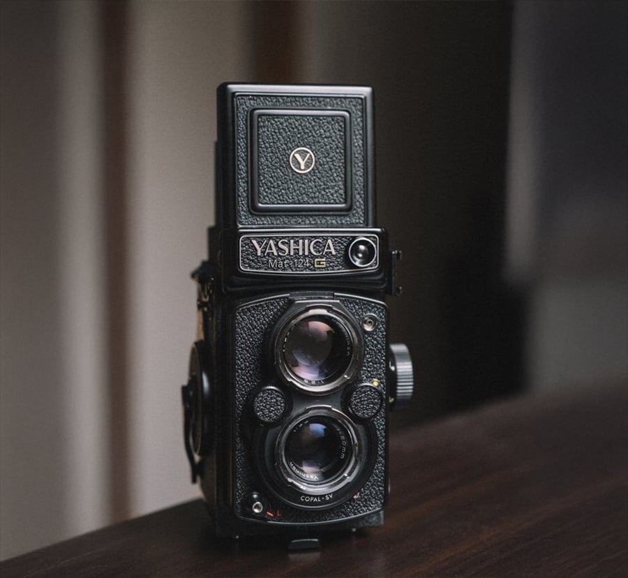 A vintage Yashica Mat-124 twin-lens reflex camera sits on a dark wooden surface, with soft natural light highlighting its textured black body and dual lenses.