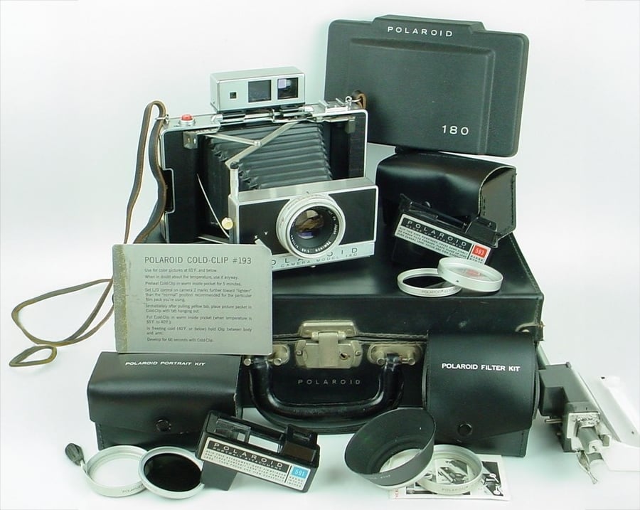 Vintage Polaroid camera with leather case, lens filters, instruction cards, and accessories arranged neatly. Items include flash units, lens kits, and labeled cases, showcasing a complete Polaroid photography set.