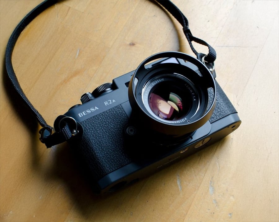 A black Bessa R2A film camera with a lens and strap rests on a wooden surface, reflecting light on its lens glass.