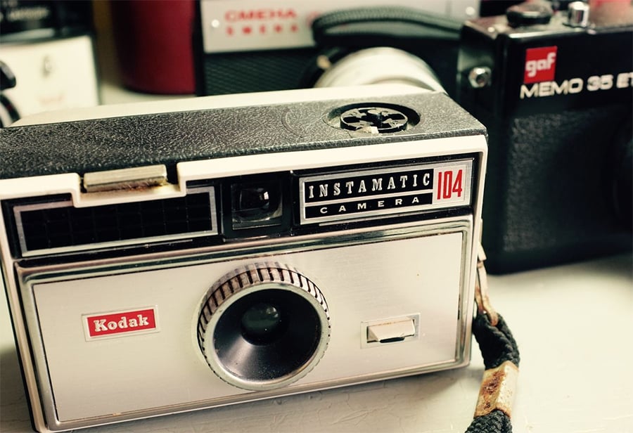 Close-up of a vintage Kodak Instamatic 104 camera, with part of a black GAF Memo 35 camera and another camera blurred in the background. The cameras show signs of age and use.