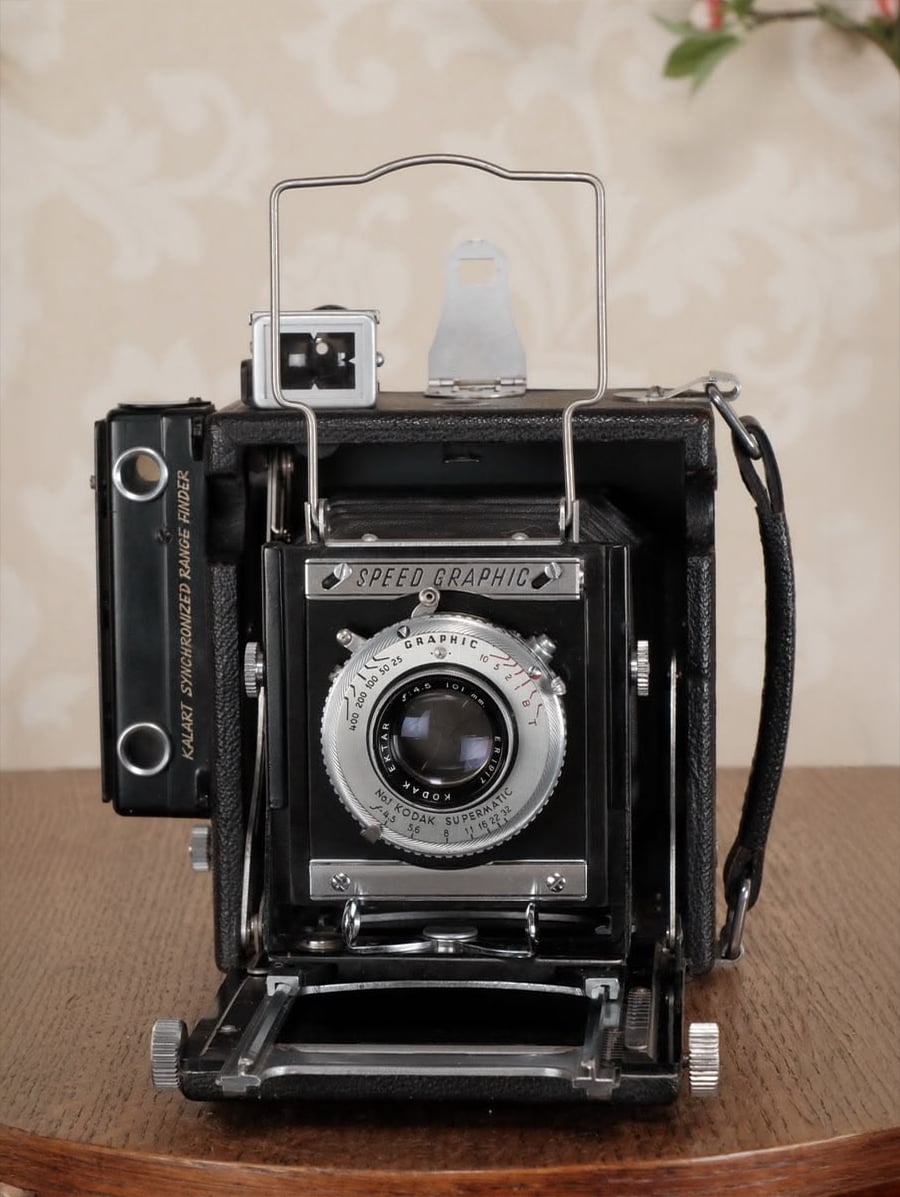 A vintage Speed Graphic press camera sits on a wooden table with a floral-patterned wallpaper in the background. The camera’s lens and metal details are visible, along with a handle and part of the film holder.