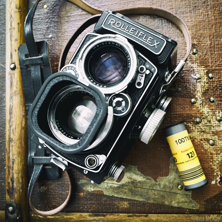 A vintage Rolleiflex twin-lens reflex camera sits on a wooden surface with a roll of Kodak 120 film beside it. A worn leather strap is visible, adding to the camera’s antique appearance.