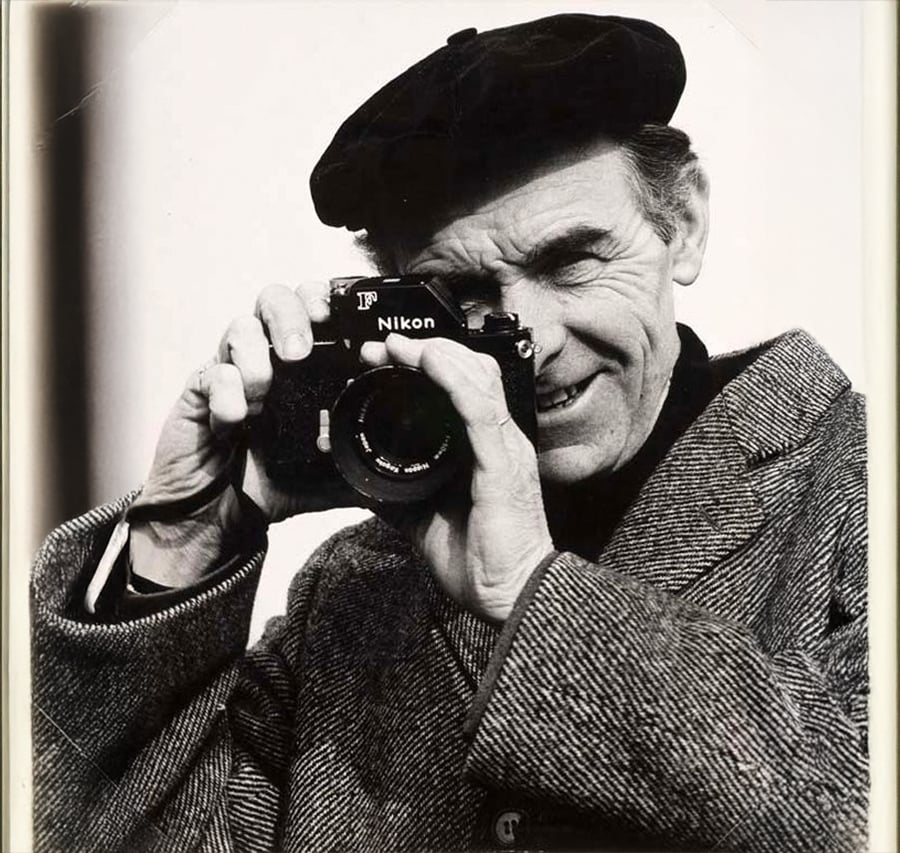 An older man wearing a beret and tweed coat smiles while holding a Nikon film camera up to his face, as if taking a picture. The image is in black and white.