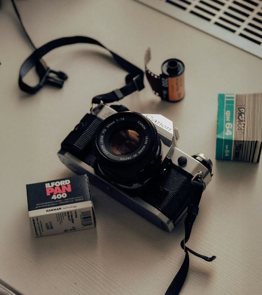 A vintage Canon film camera sits on a table with a lens attached. Nearby are a roll of exposed film, a box of Ilford Pan 400 film, and a pack of Fujicolor film. A camera strap is also visible.